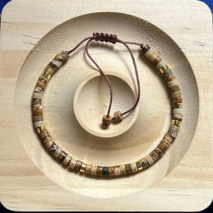 May include: A brown, beige, and gold beaded bracelet with a brown cord closure. The bracelet is made of natural stone beads and is displayed on a wooden surface.