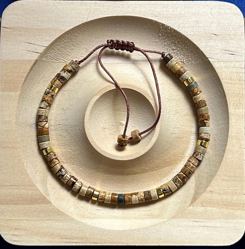 May include: A brown, beige, and gold beaded bracelet with a brown cord closure. The bracelet is made of natural stone beads and is displayed on a wooden surface.
