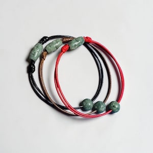 Può includere: Tre braccialetti in cordino regolabile nei colori nero, marrone e rosso. Ogni braccialetto presenta una singola perla in pietra verde intagliata di forma cilindrica con una finitura liscia. I cordini sono fissati con nodi scorrevoli regolabili.