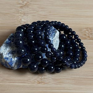 Pode incluir: Um conjunto de pulseiras de contas azuis escuras feitas de pedra de areia azul. As pulseiras estão sobre uma superfície de madeira marrom claro.