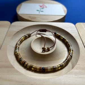 May include: A brown and gold beaded bracelet with a brown cord closure. The bracelet is displayed on a wooden surface.