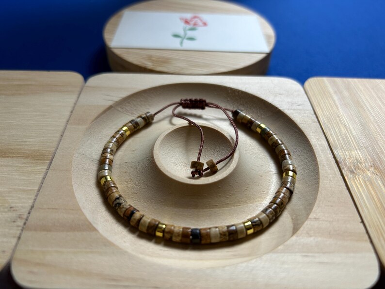 May include: A brown and gold beaded bracelet with a brown cord closure. The bracelet is displayed on a wooden surface.