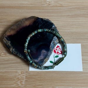 May include: A green and gold beaded bracelet with a green cord closure. The bracelet is lying on a piece of agate with a dark purple and black pattern. A white card with a red rose illustration is behind the bracelet.