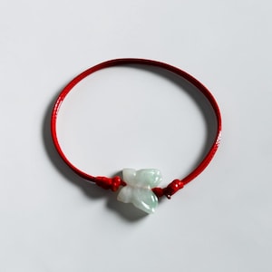 May include: A bracelet featuring a smooth, pale green, translucent stone carved into the shape of a butterfly. The stone is centered on a thin, vibrant red cord that is secured with small, knotted red accents on either side of the butterfly.