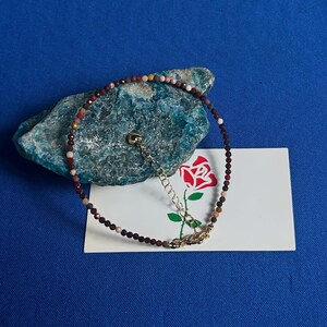 Op de afbeelding: Een delicate kralen armband met een gouden sluiting. De kralen zijn een mix van rood, bruin en wit, en de armband ligt op een groene steen.