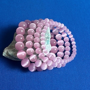 以下が含まれることがあります： 丸いビーズが付いたピンクの宝石のブレスレット4本セット。ブレスレットは重ねて、ざらざらした灰色の岩の上に置かれています。