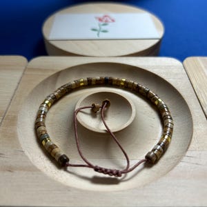 May include: A brown, beige, and gold beaded bracelet with a brown cord closure. The bracelet is displayed on a wooden surface.