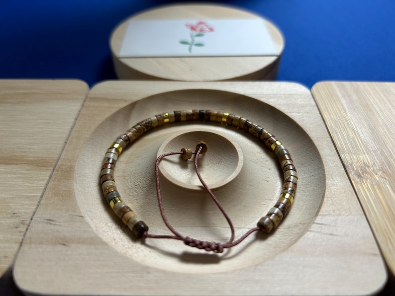 May include: A brown, beige, and gold beaded bracelet with a brown cord closure. The bracelet is displayed on a wooden surface.
