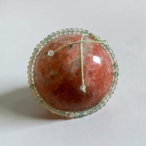 May include: A smooth, spherical orange and red stone orb is encircled by a delicate, thin string of small, translucent green beads. The green beaded strand is tied at the top with a light-colored cord, creating a decorative, layered appearance.