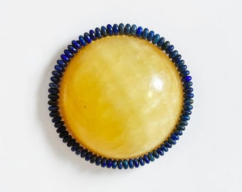 May include: A round, yellow gemstone is encircled by a beaded, dark blue border. The gemstone has a smooth, polished surface. The dark blue beads are small and evenly spaced, creating a contrasting frame against the yellow center.