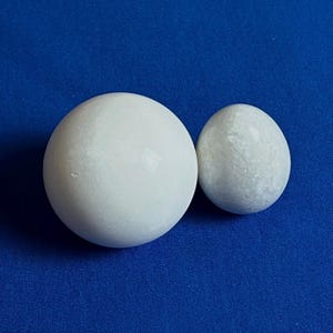 Puede incluir: Dos esferas de cerámica blanca, una más grande que la otra, sobre un fondo azul. La esfera más grande es lisa, mientras que la esfera más pequeña tiene una superficie texturizada.