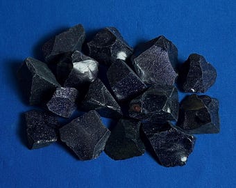 Puede incluir: Un montón de virutas de cristal de obsidiana negra con un brillo brillante e iridiscente. Las virutas son de varios tamaños y formas, y están dispuestas sobre un fondo azul.