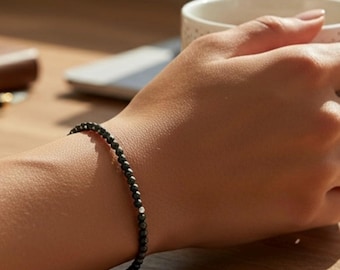 May include: A fine black beaded bracelet with a single silver bead. The bracelet is displayed on a wrist, with a wooden surface in the background. The bracelet is a simple, elegant piece of jewellery.
