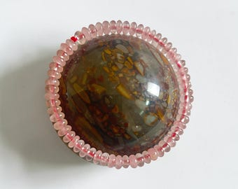 May include: A round, polished stone with a mix of brown, yellow, and gray patterns, encircled by a strand of small, pink beads. The stone has a glossy finish, reflecting light.
