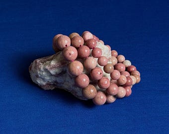 May include: A strand of pink and white gemstone beads, likely made of rhodonite, arranged in a circular shape on a piece of rough, pink and white stone.