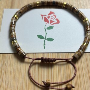 May include: A brown and gold beaded bracelet with a brown cord closure. The beads are made of natural stone and have a speckled pattern.