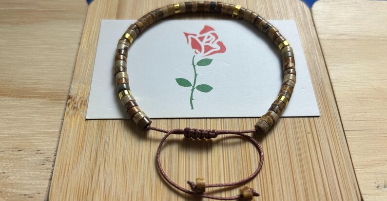 May include: A brown and gold beaded bracelet with a brown cord closure. The beads are made of natural stone and have a speckled pattern.