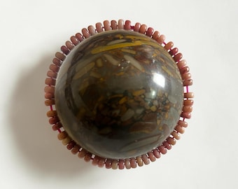 May include: A polished, spherical stone with a mottled pattern of brown, gray, and yellow hues. The stone is encircled by a ring of small, reddish-brown beads, creating a decorative border. The overall design is centered on a white background.
