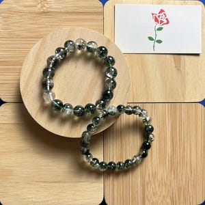 May include: Two green and clear crystal beaded bracelets. The bracelets are made of round beads with a speckled green and clear design. The bracelets are on a wooden surface.