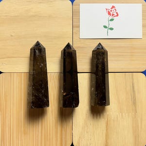 May include: Three smoky quartz crystal points on a wooden surface. The points are all about the same size and shape, and they have a dark brown colour.