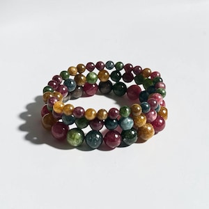 May include: Three beaded bracelets in shades of red, green, yellow, and blue. The bracelets are made of round, polished beads and are stacked on top of each other. The bracelets are on a white background.