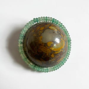 May include: A polished, spherical gemstone with a mottled pattern of brown, yellow, and gray. A ring of small, light green beads encircles the stone. The gemstone is set against a white background.