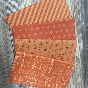 May include: Five fabric swatches in various shades of orange and brown with different patterns. The patterns include a plaid, a small cross pattern, a floral pattern, and two patterns with text. The text includes the words "apple pick", "orchard", "mums", "ring season", "trees", "cider", "baking", "fall", "yummy", and "patch".