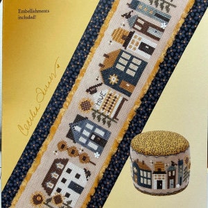 May include: A cross stitch pattern featuring a row of houses with sunflowers and the text "HIVE". The pattern is on a beige fabric with a black and yellow border. The pattern is also shown on a small, round, yellow and black fabric cushion.