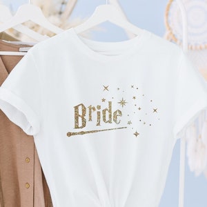 Op de afbeelding: Wit T-shirt met een gouden glitterontwerp met de tekst "Bride" met een toverstaf en sterren. Het shirt hangt aan een witte hanger.