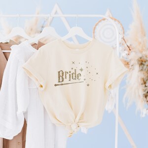 May include: A cream-colored t-shirt with the word "Bride" written in gold glitter with a wand and stars. The shirt is hanging on a white hanger.