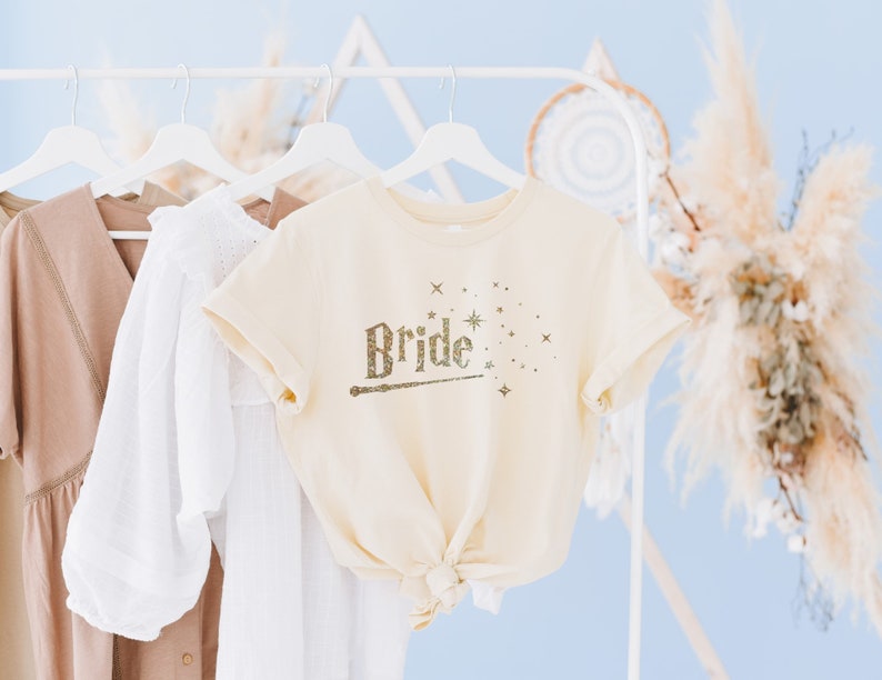 May include: A cream-colored t-shirt with the word "Bride" written in gold glitter with a wand and stars. The shirt is hanging on a white hanger.