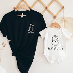 May include: A black t-shirt featuring a white line-art dinosaur and the text DADDYSAURUS, paired with a white baby bodysuit featuring a black line-art dinosaur hatching from an egg and the text BABYSAURUS, both displayed on wooden hangers.