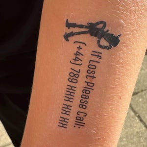 May include: A black temporary tattoo featuring a silhouette of a small character with a hat and the text "If Lost Please Call: (+44) 789 XXX XX XX" printed in a sans-serif font. The design is applied directly to the skin with a clear adhesive border.