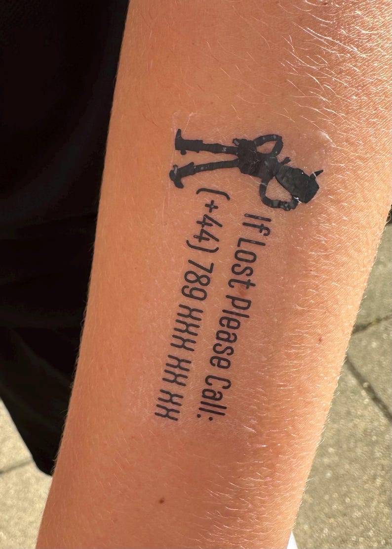 May include: A black temporary tattoo featuring a silhouette of a small character with a hat and the text "If Lost Please Call: (+44) 789 XXX XX XX" printed in a sans-serif font. The design is applied directly to the skin with a clear adhesive border.
