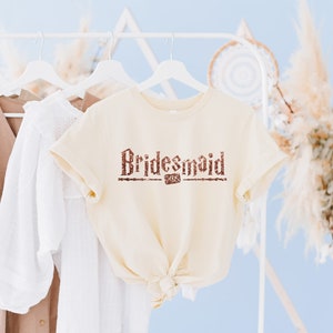 Op de afbeelding: Een cr&egrave;mekleurig T-shirt met het woord "Bridesmaid" in bruine glitterletters op de voorkant.