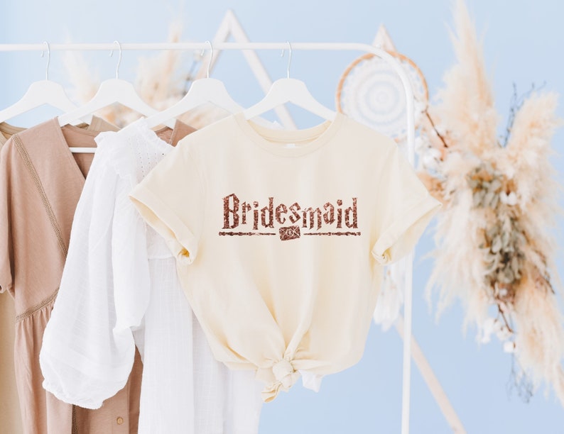 Op de afbeelding: Een cr&egrave;mekleurig T-shirt met het woord "Bridesmaid" in bruine glitterletters op de voorkant.