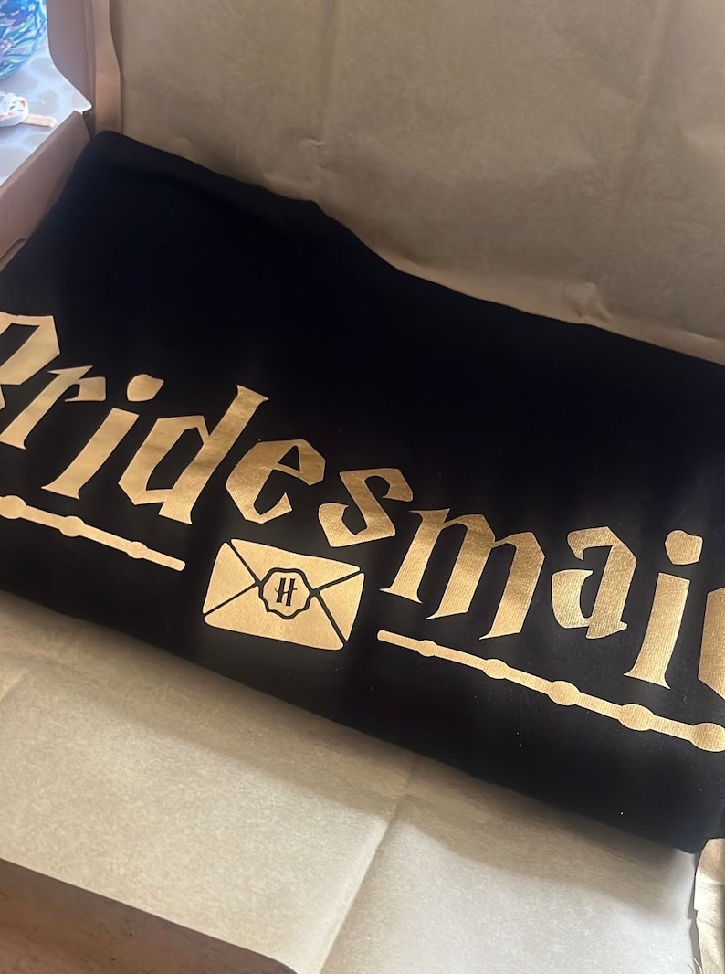 May include: Black t-shirt with gold lettering that says "Bridesmaid" and a gold envelope with a "H" inside.
