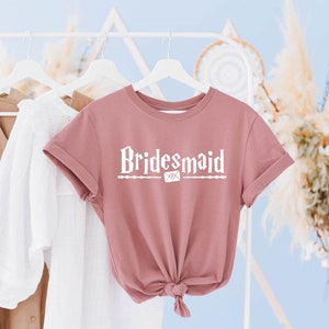 May include: A dusty rose t-shirt with the text "Bridemaid" in white lettering. The text is stylized with a wand and an envelope graphic.