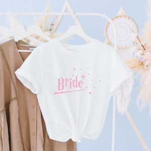 Op de afbeelding: Wit T-shirt met een roze afbeelding van een toverstaf en het woord "Bride" erop. Het shirt hangt aan een witte hanger.
