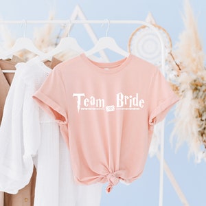 Op de afbeelding: Een roze T-shirt met de tekst "Team Bride" in witte letters. De tekst is gestyled als een toverstaf uit de Harry Potter-serie.