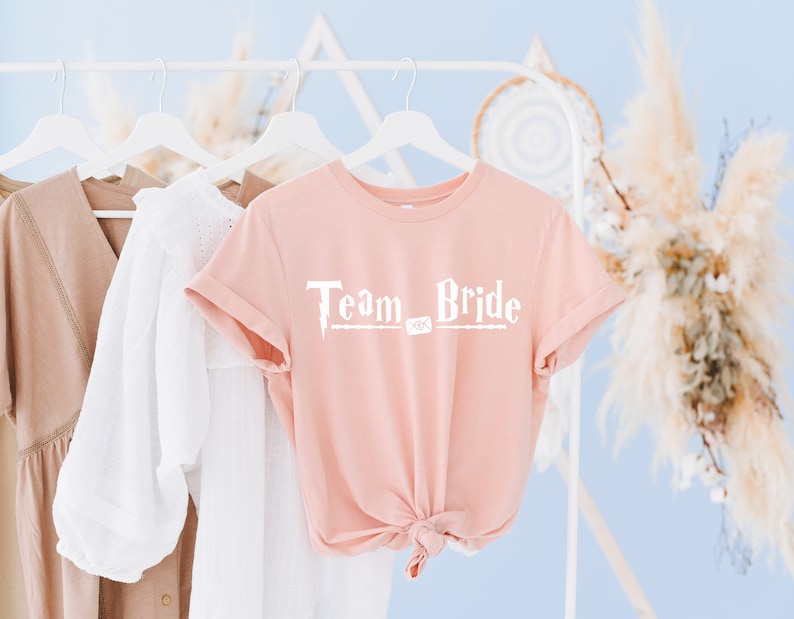 Op de afbeelding: Een roze T-shirt met de tekst "Team Bride" in witte letters. De tekst is gestyled als een toverstaf uit de Harry Potter-serie.