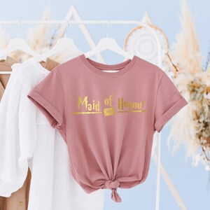 Op de afbeelding: Een roze T-shirt met de tekst "Maid of Honour" in gouden letters en een toverstafontwerp.