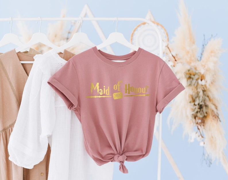 Op de afbeelding: Een roze T-shirt met de tekst "Maid of Honour" in gouden letters en een toverstafontwerp.