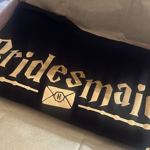 Op de afbeelding: Zwart T-shirt met gouden letters die "Bridesmaid" zeggen en een gouden envelop met een "H" erin.