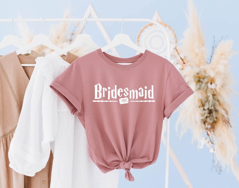 Op de afbeelding: Een stoffig roze t-shirt met de tekst "Bridemaid" in witte letters. De tekst is gestyled in de stijl van een Harry Potter-ontwerp met een witte envelop en een toverstaf.