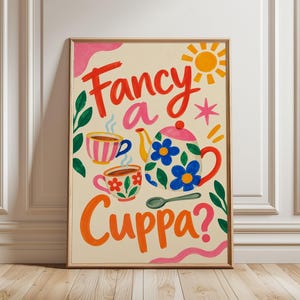 Puede incluir: Una lámina artística enmarcada sobre fondo crema con el texto Fancy a Cuppa? en rojo y naranja. La ilustración incluye una tetera blanca con flores azules, dos tazas de té estampadas con vapor, una cuchara verde, un sol amarillo y detalles rosas.