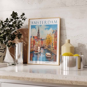 May include: A framed vintage poster with a colourful illustration of Amsterdam, Netherlands. The poster has the text "AMSTERDAM" in a gold frame. The poster is on a white countertop with a silver jug, a wooden chopping board, a silver bowl with lemons, and a tan ceramic vase.