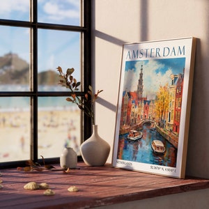 May include: A framed vintage travel poster with a colourful illustration of Amsterdam, Netherlands. The poster features a canal with boats, buildings, and a tall church steeple. The text on the poster reads "AMSTERDAM, NETHERLANDS, 52.3678&deg; N, 4.9041&deg; E".