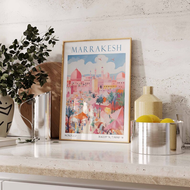 May include: A vintage travel poster with a gold frame depicting a colorful scene of Marrakech, Morocco. The poster features a pink cityscape with palm trees and people in the foreground. The text "MARRAKESH" is printed in bold letters at the top of the poster.