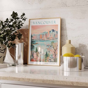May include: A vintage-style poster with a gold frame depicting a cityscape of Vancouver, Canada. The poster has a pink and blue color scheme and features the text "Vancouver" and "Canada" along with the coordinates "49.2827&deg; N, 123.1207&deg; W".